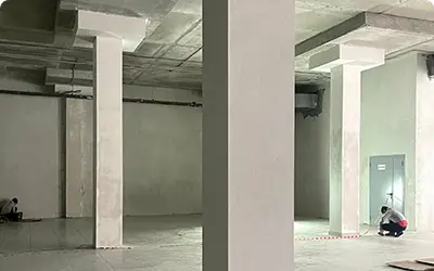 Ход профессиональной отделки коммерческого помещения: штукатурка стен, колонны и стяжка пола. Внутренний вид большого коммерческого помещения с оштукатуренными колоннами и стенами, где рабочий на полу проводит отделочные работы.